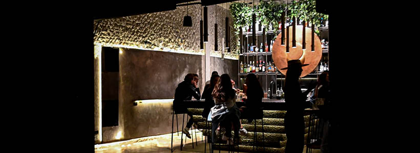 Clienti al bancone del Bar Tana, immersi in un’atmosfera intima con illuminazione soffusa e materiali grezzi.