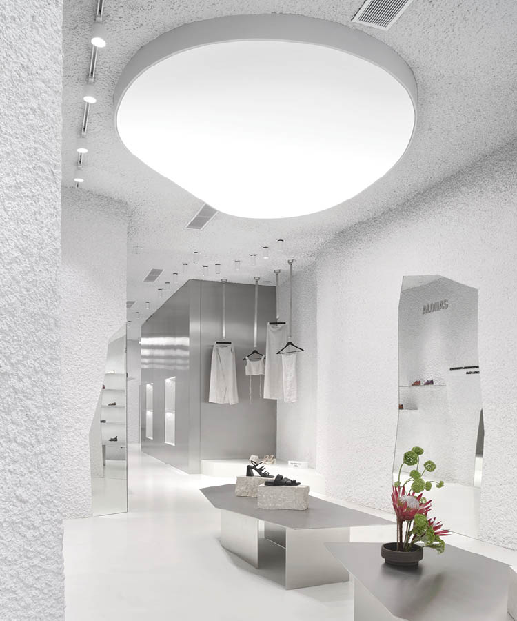 Interior of the Alohas flagship store in Barcelona featuring textured white walls, a large organic-shaped ceiling light, satin metal volumes, and display tables with footwear