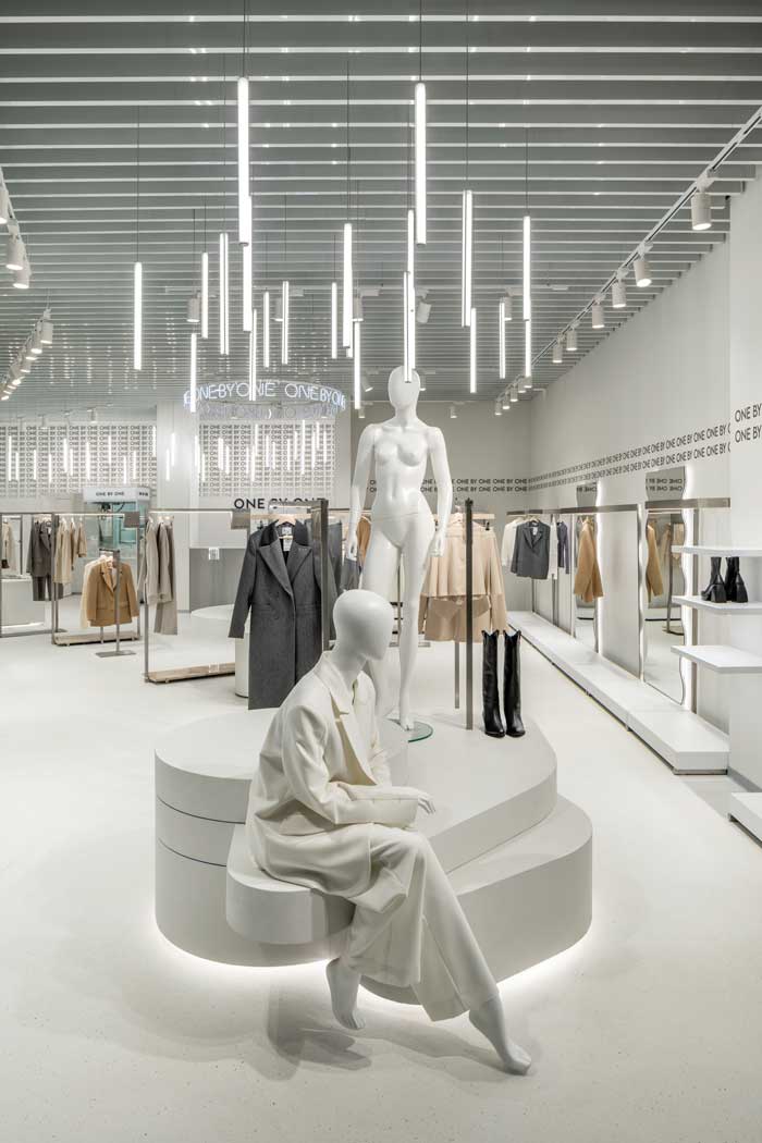 Area vendita dello showroom One By One con illuminazione a sospensione lineare, pedana circolare con manichini e rack di abbigliamento