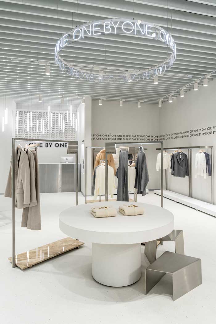 Tavolo espositivo rotondo e insegna al neon circolare nello showroom One By One, con espositori minimal e capi appesi