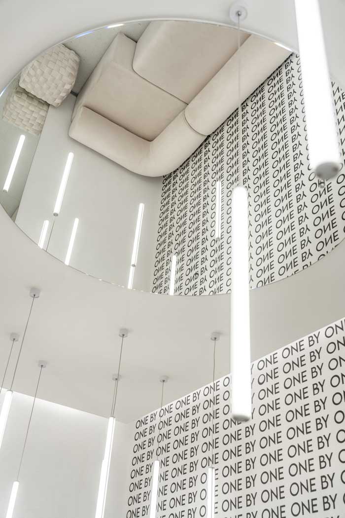 Dettaglio del soffitto nello showroom One By One con lampade tubolari sospese, specchio circolare e grafica tipografica a parete