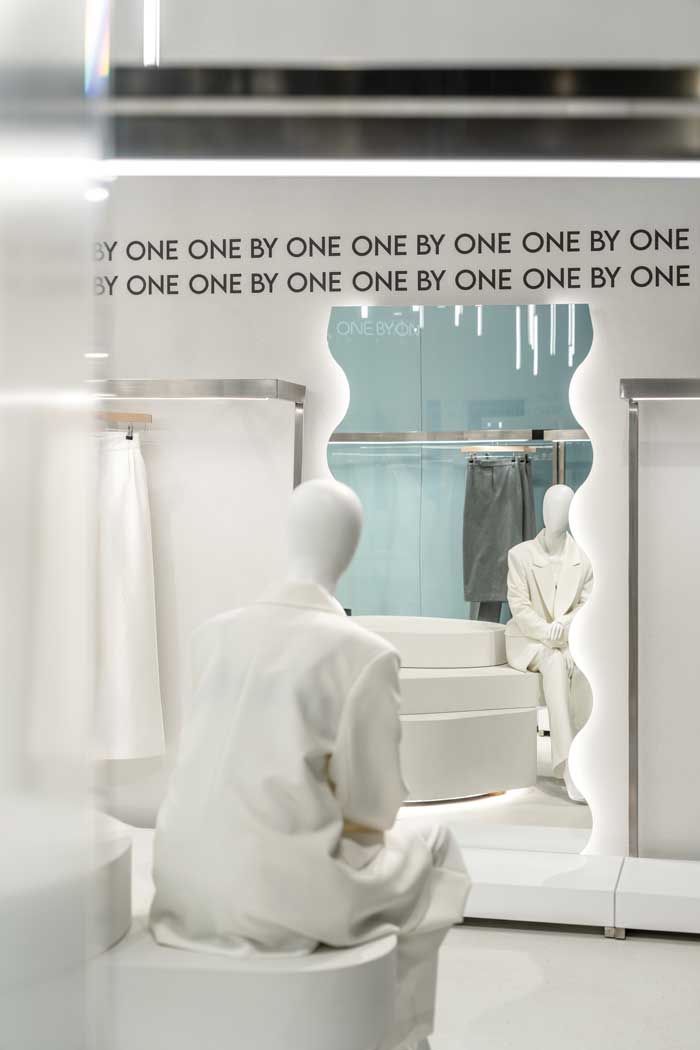 Manichini e specchio sagomato nello showroom One By One, con grafica tipografica ripetuta e allestimento minimale
