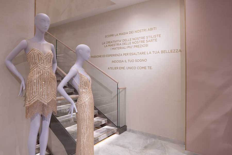Interno elegante del nuovo Atelier Emé a Palazzo Pertusati in Via della Spiga, Milano: due manichini con abiti dorati di alta moda davanti a una scala di marmo fior di pesco, illuminazione raffinata e dettagli di design contemporaneo
