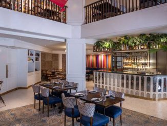 Interno di un ristorante in stile Art Déco con bar frontale decorato, piante sospese, sedie imbottite e pavimento con piastrelle a motivi geometrici. Un balcone in legno al piano superiore aggiunge ulteriore spazio per la ristorazione.