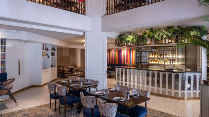 Interno di un ristorante in stile Art Déco con bar frontale decorato, piante sospese, sedie imbottite e pavimento con piastrelle a motivi geometrici. Un balcone in legno al piano superiore aggiunge ulteriore spazio per la ristorazione.