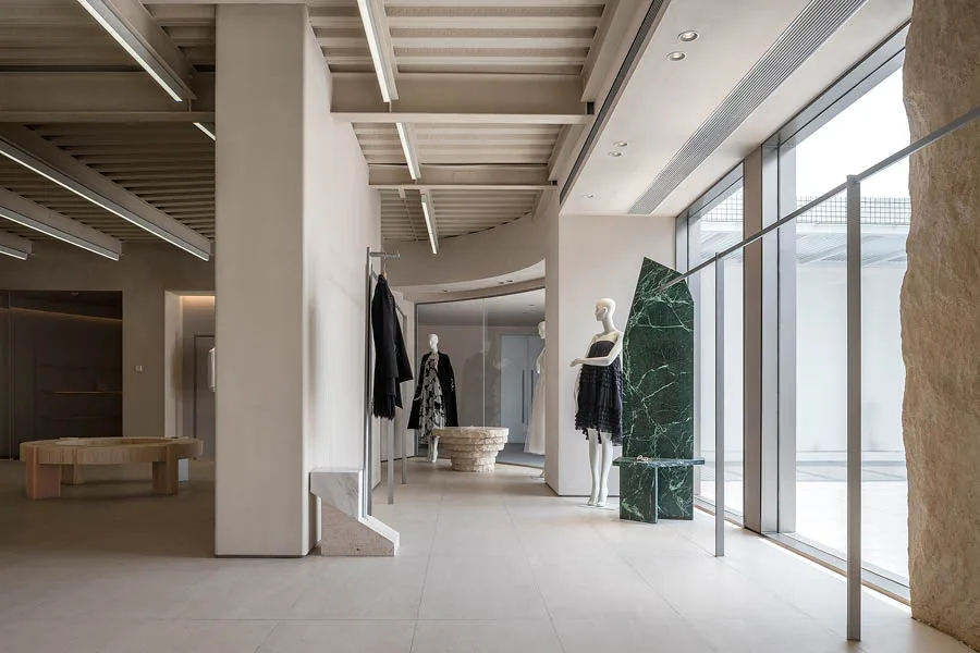 Interno di un concept store con design minimalista, arredi moderni e utilizzo di materiali naturali come marmo e pietra. Manichini esposti con abbigliamento elegante e grandi vetrate che offrono luminosità all'ambiente