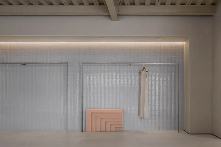 Dettaglio di un concept store con parete rivestita in piastrelle minimaliste, un appendiabiti in metallo con un abito bianco appeso e un elemento decorativo geometrico in tonalità rosa