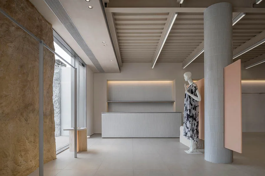Interno di un concept store con design minimalista, pareti in texture naturali, un bancone moderno, un manichino con abito floreale e ampie vetrate che illuminano l'ambiente.