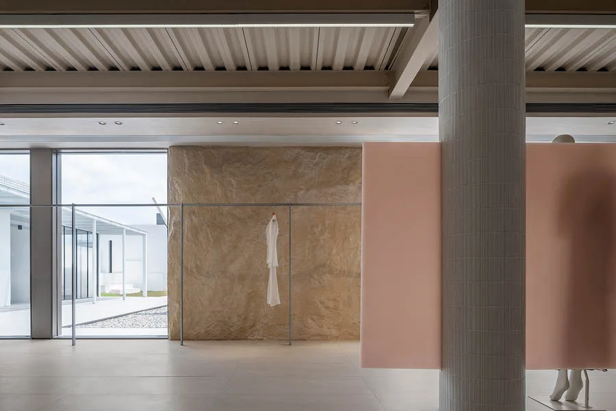 Interno di un concept store con pareti in texture naturali, divisori rosa pastello, una colonna rivestita e un capo bianco appeso su una struttura minimalista in metallo. Grandi vetrate illuminano l'ambiente con vista sull'esterno