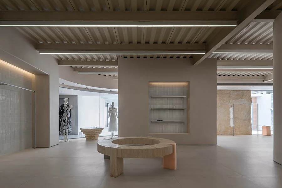 Interno di un concept store con arredi minimalisti, tavolo circolare in materiale naturale, espositori integrati nella parete e manichini con abbigliamento elegante. Il design moderno è caratterizzato da tonalità neutre e linee pulite