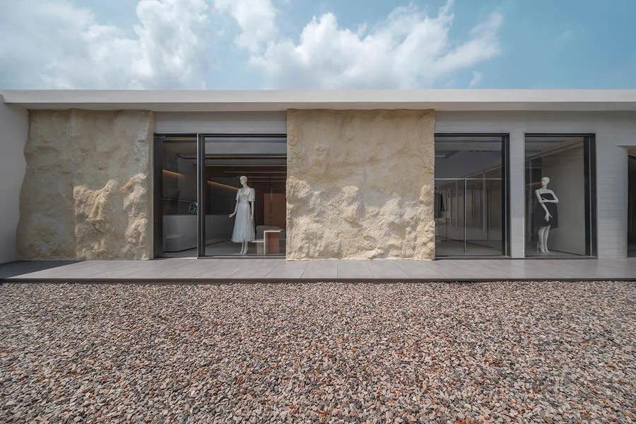 Esterno di un concept store con pareti in texture naturale, ampie vetrate che mostrano l'interno con manichini esposti, pavimentazione in ghiaia e cielo azzurro con nuvole