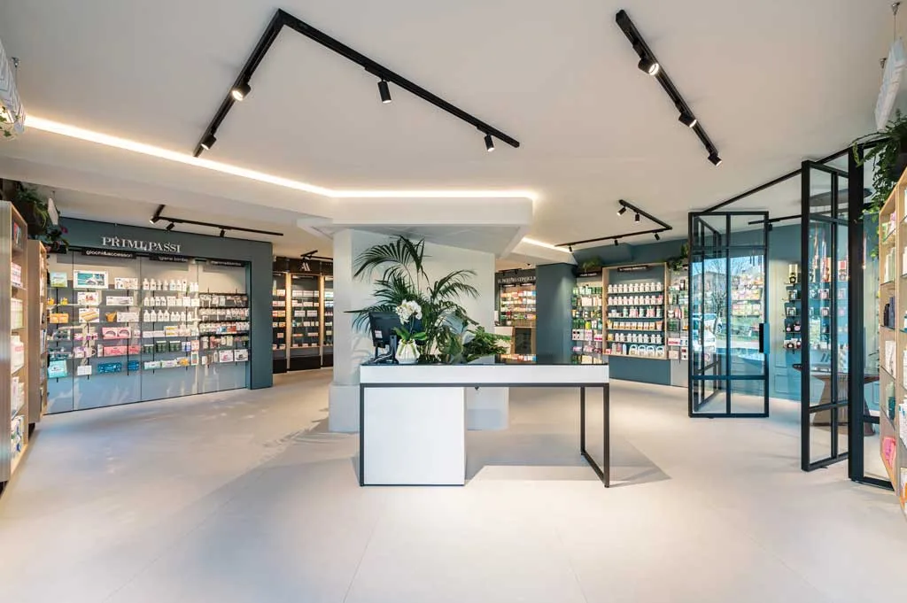 Interno di una farmacia moderna con arredi minimalisti, illuminazione a LED e scaffali ben organizzati con prodotti in esposizione. Un banco accoglienza con piante verdi dona un tocco naturale allo spazio.