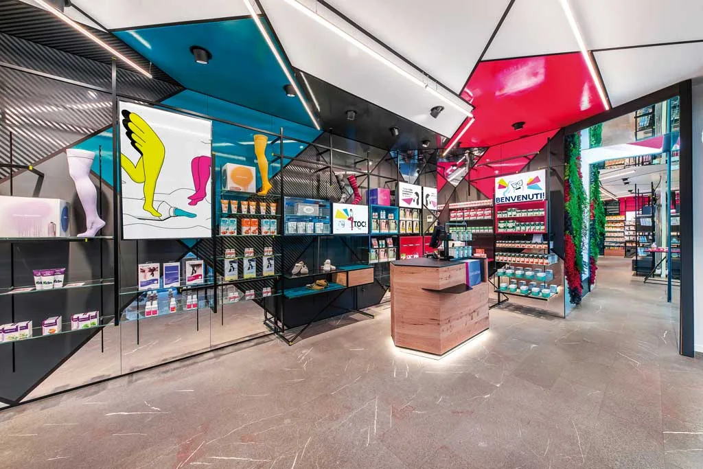 Design innovativo di una farmacia con elementi geometrici colorati sul soffitto, pareti con espositori in vetro e un banco cassa in legno. Illuminazione dinamica e decorazioni vivaci creano un ambiente contemporaneo.