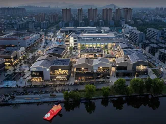 Vista aerea notturna di SHAOXING PLACE by MixC, un complesso urbano riqualificato che combina architettura industriale e retail moderno, affacciato su un canale con un'installazione galleggiante.