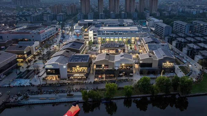Vista aerea notturna di SHAOXING PLACE by MixC, un complesso urbano riqualificato che combina architettura industriale e retail moderno, affacciato su un canale con un'installazione galleggiante.
