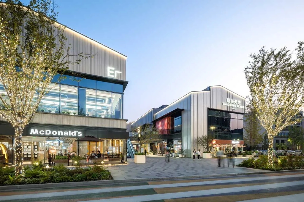 Area commerciale di SHAOXING PLACE by MixC al tramonto, con edifici moderni dal design industriale, illuminazioni decorative e spazi retail, tra cui un McDonald's, immersi in un ambiente urbano dinamico.