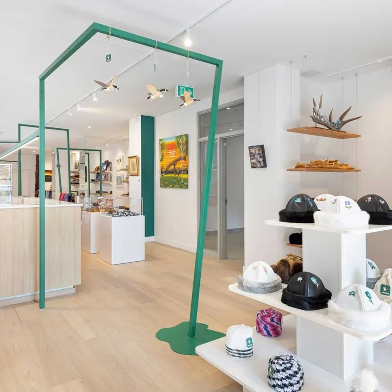 Interior of the Wachiya Flagship Store with a modern and minimalist design, featuring suspended green geometric structures that enhance the space. In the foreground, a display of fur hats with traditional Cree embroidery is arranged on white shelves. In the background, wooden shelves showcase handcrafted items, including antler sculptures and wood carvings. The white walls are adorned with colorful paintings, while a light wood counter and display cases complete the setting, emphasizing the fusion of Indigenous tradition and contemporary retail design.
