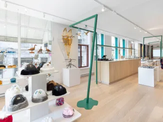 Interno luminoso e moderno del Wachiya Flagship Store, con un design minimalista che valorizza l'artigianato Cree. L'allestimento include espositori bianchi con cappelli in pelliccia e accessori tradizionali, strutture metalliche verdi dal design contemporaneo e un ampio bancone in legno naturale. Le grandi finestre permettono alla luce naturale di entrare, creando un ambiente accogliente e raffinato. Sullo sfondo, scaffali con sculture di uccelli in legno e ciaspole tradizionali arricchiscono l’atmosfera culturale del negozio.