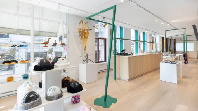 Interno luminoso e moderno del Wachiya Flagship Store, con un design minimalista che valorizza l'artigianato Cree. L'allestimento include espositori bianchi con cappelli in pelliccia e accessori tradizionali, strutture metalliche verdi dal design contemporaneo e un ampio bancone in legno naturale. Le grandi finestre permettono alla luce naturale di entrare, creando un ambiente accogliente e raffinato. Sullo sfondo, scaffali con sculture di uccelli in legno e ciaspole tradizionali arricchiscono l’atmosfera culturale del negozio.