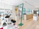 Interno luminoso e moderno del Wachiya Flagship Store, con un design minimalista che valorizza l'artigianato Cree. L'allestimento include espositori bianchi con cappelli in pelliccia e accessori tradizionali, strutture metalliche verdi dal design contemporaneo e un ampio bancone in legno naturale. Le grandi finestre permettono alla luce naturale di entrare, creando un ambiente accogliente e raffinato. Sullo sfondo, scaffali con sculture di uccelli in legno e ciaspole tradizionali arricchiscono l’atmosfera culturale del negozio.