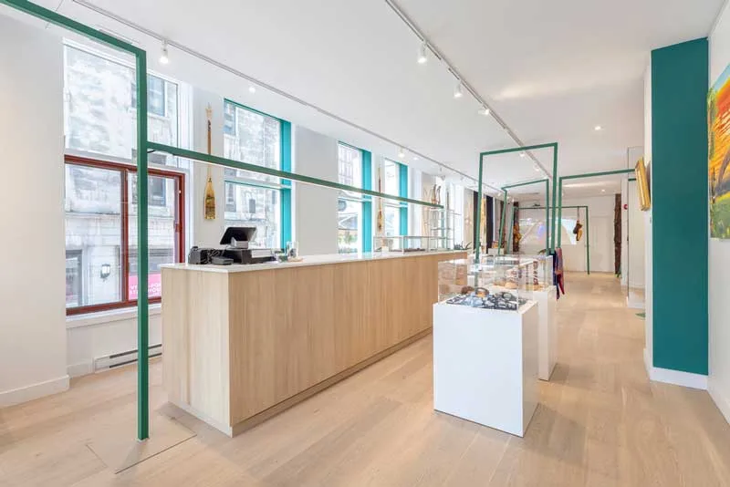 Interior of the Wachiya Flagship Store with a modern and minimalist design that highlights Cree craftsmanship. The space features a large light wood counter, framed by green metal structures that create a dynamic visual effect. The large windows with turquoise details allow natural light to illuminate the space, while white pedestals showcase jewelry and handcrafted items. Traditional elements, such as snowshoes and wood carvings, adorn the walls, emphasizing the fusion of Indigenous culture and contemporary retail design.