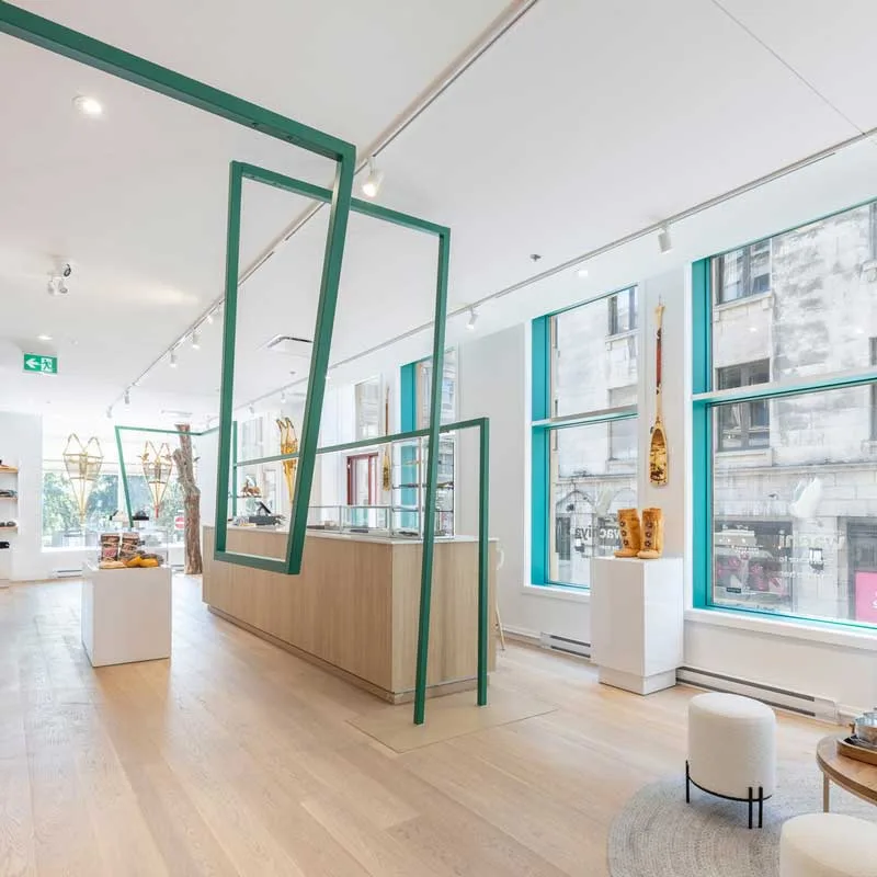 Interior of the Wachiya Flagship Store with a minimalist and contemporary design, featuring large windows framed by turquoise elements that illuminate the space with natural light. Suspended green geometric structures create a dynamic effect, while the light wood counter adds warmth to the environment. Cree craftsmanship elements, such as traditional snowshoes and wooden sculptures, are displayed on white pedestals, emphasizing the fusion of tradition and modernity in the store's retail design.