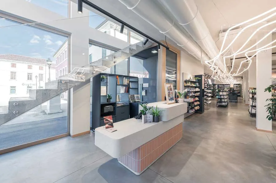 Area accoglienza della Farmacia Fecchio progettata da AMlab, con bancone moderno, scala in vetro e metallo, grandi vetrate e illuminazione LED decorativa a soffitto