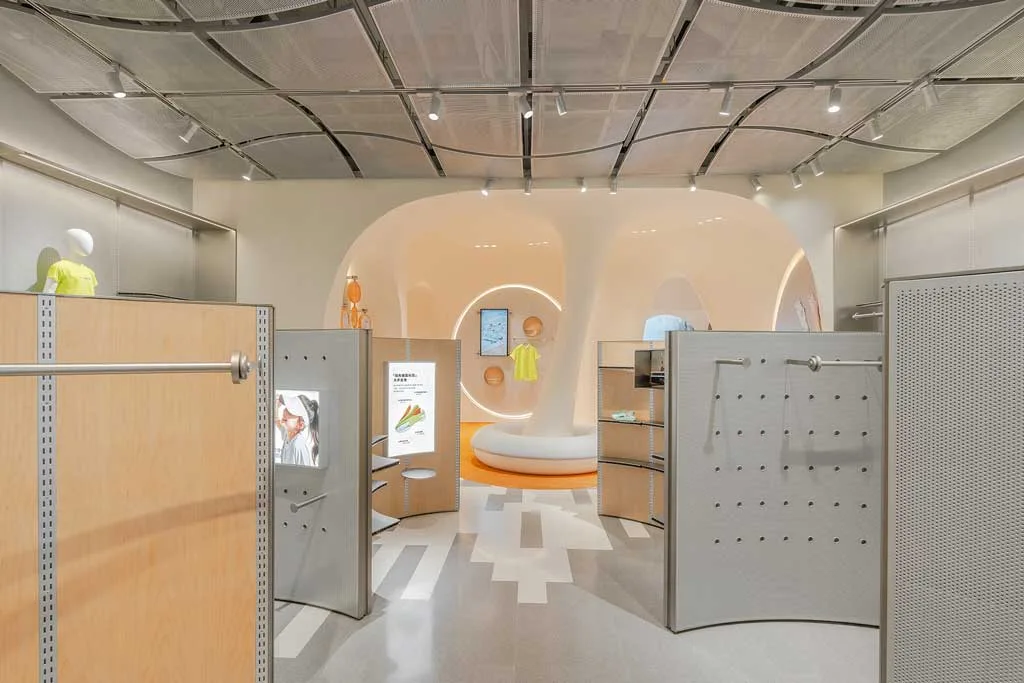 Vista interna del negozio Moodytiger con arredi curvi in legno chiaro, pannelli espositivi metallici forati e area centrale con pareti organiche retroilluminate e display digitali per l’abbigliamento tecnico per bambini.
