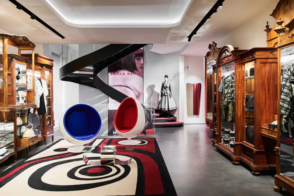 ephemeral-di-antonio-marras-boutique-temporanea-via-montenapoleone-milano_1024x683 Zona inferiore della boutique temporanea Ephemeral di Antonio Marras a Milano, con scala elicoidale nera, arredi vintage in legno e poltrone di design, durante la Milan Design Week 2025.