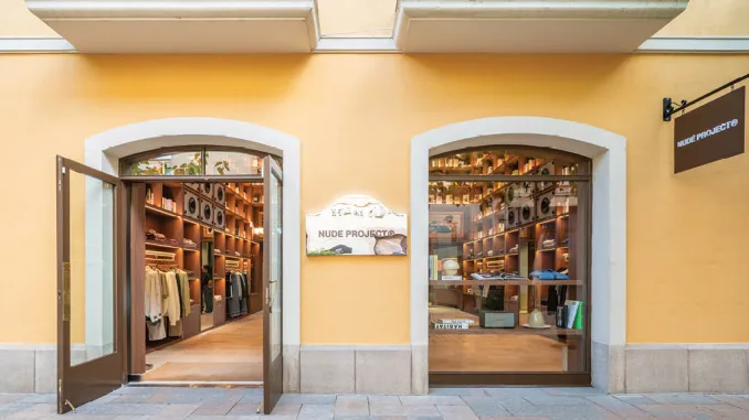 Facciata del flagship store Nude Project a La Roca Village con ingresso vetrato e libreria in legno visibile all'interno, parte del concept "Nude Library".