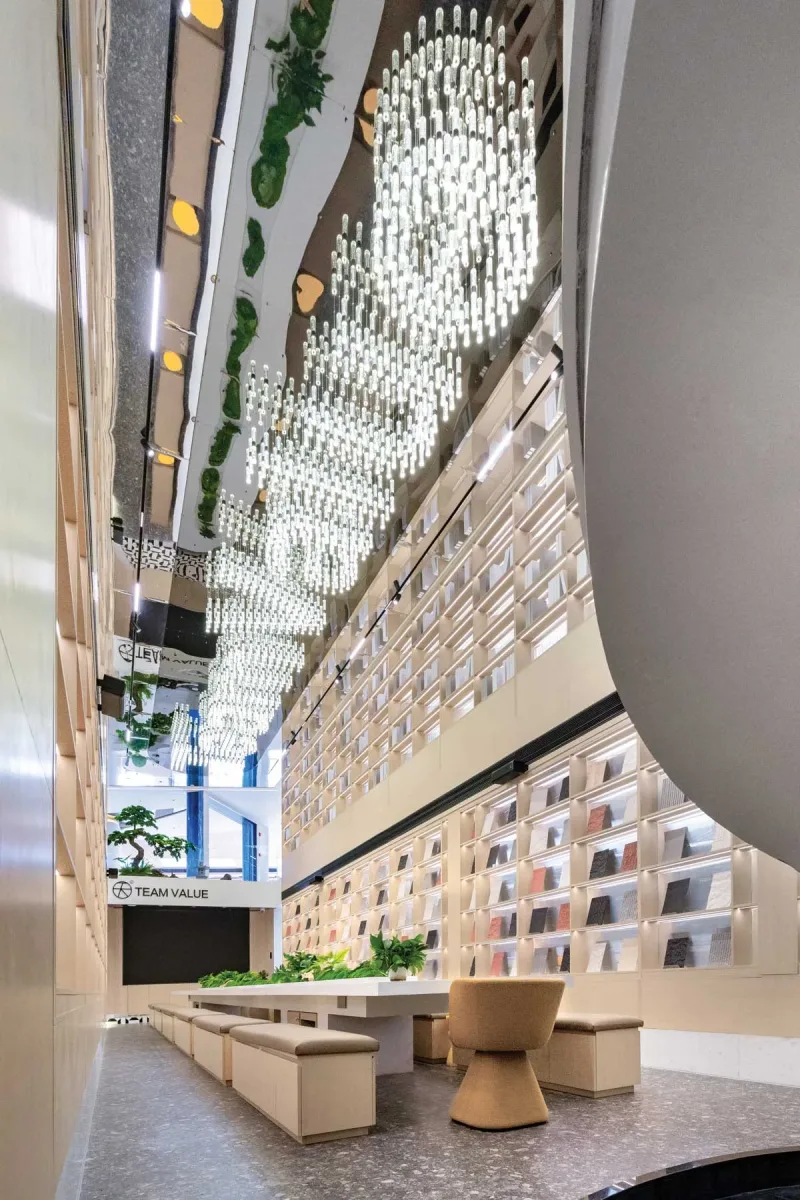 Tavolo centrale circondato da sedute nello showroom Team Value, con pareti espositive di campioni materiali e soffitto a specchio con installazione luminosa sospesa