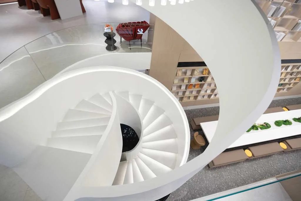 White spiral staircase viewed from above inside the Team Value showroom, with meeting table and bookshelf wall in the background