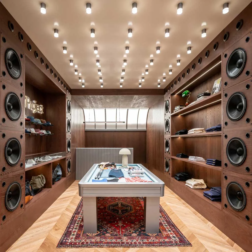 Sala “Shining” del flagship store Nude Project con esposizione centrale ispirata a un tavolo da biliardo, pannelli in legno e impianto audio integrato