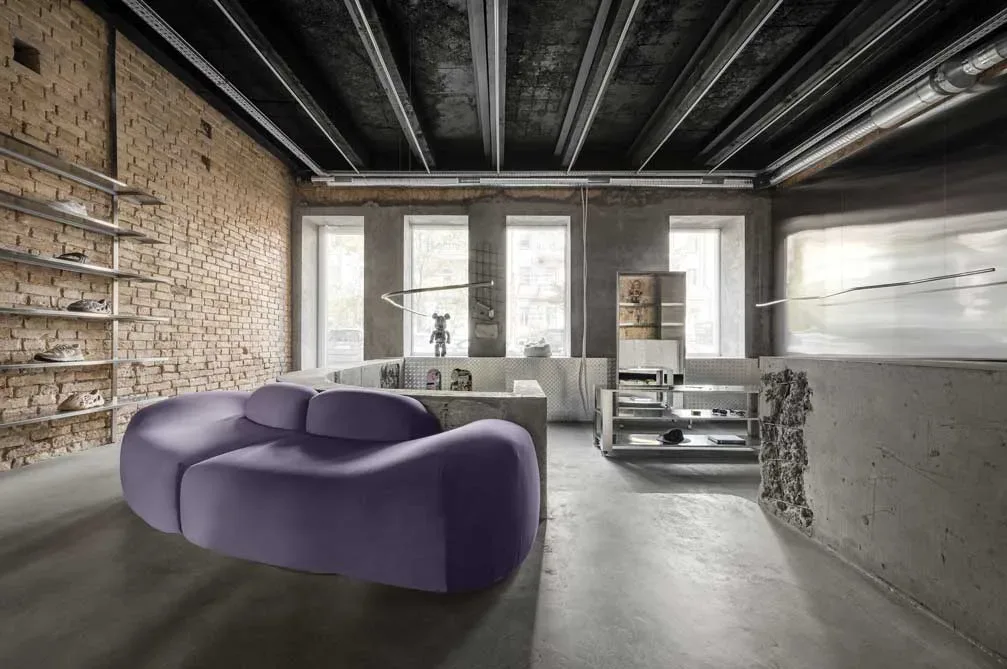 Vista dell’area lounge del concept store Storage X Domaine 17 a Kiev, con divano viola dalle forme fluide, pareti in mattoni e arredi industriali.