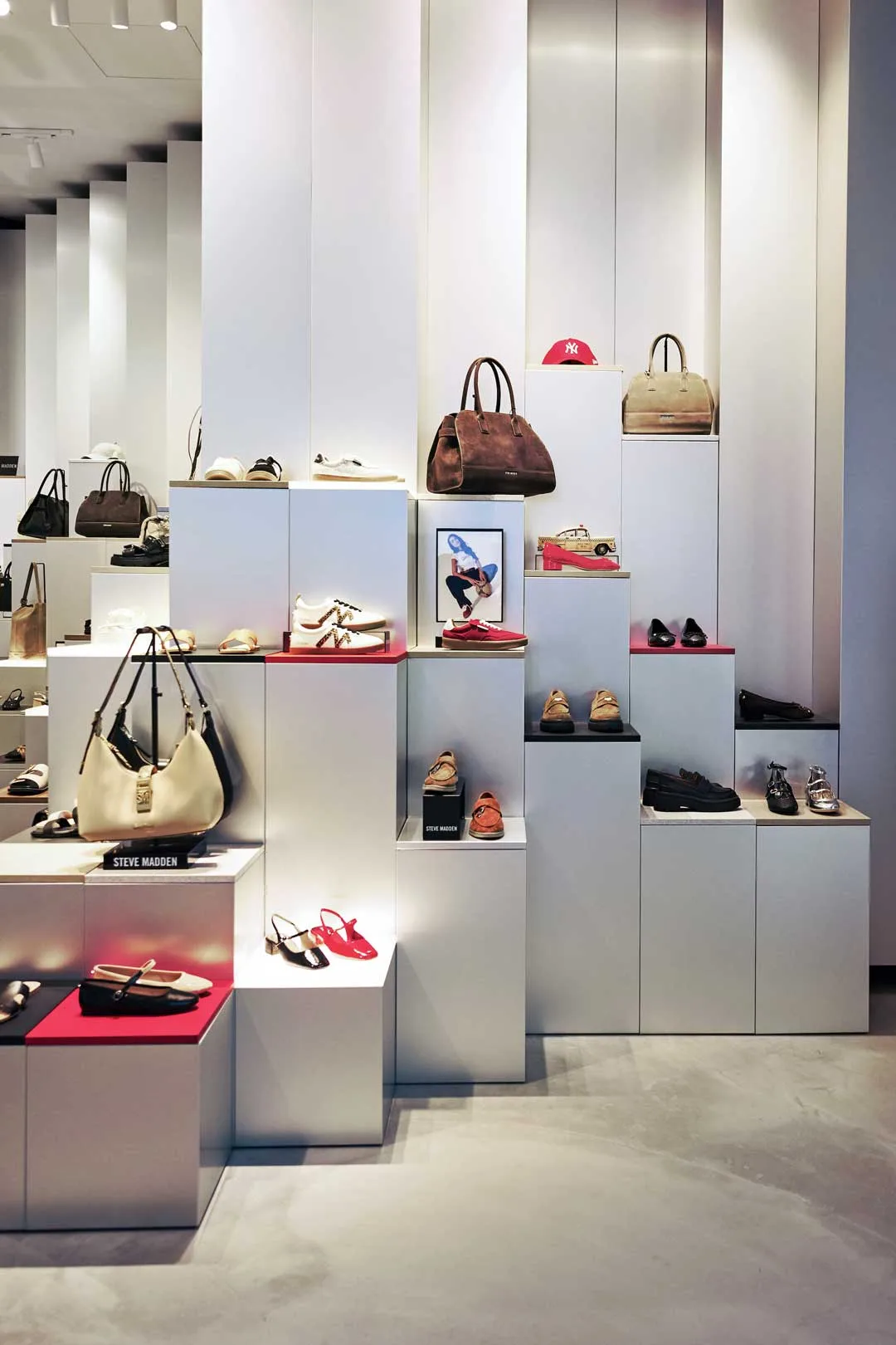Display del concept store Steve Madden nei Paesi Bassi con pedane modulari in tonalità neutre e dettagli in rosso, utilizzate per l’esposizione di borse e calzature.