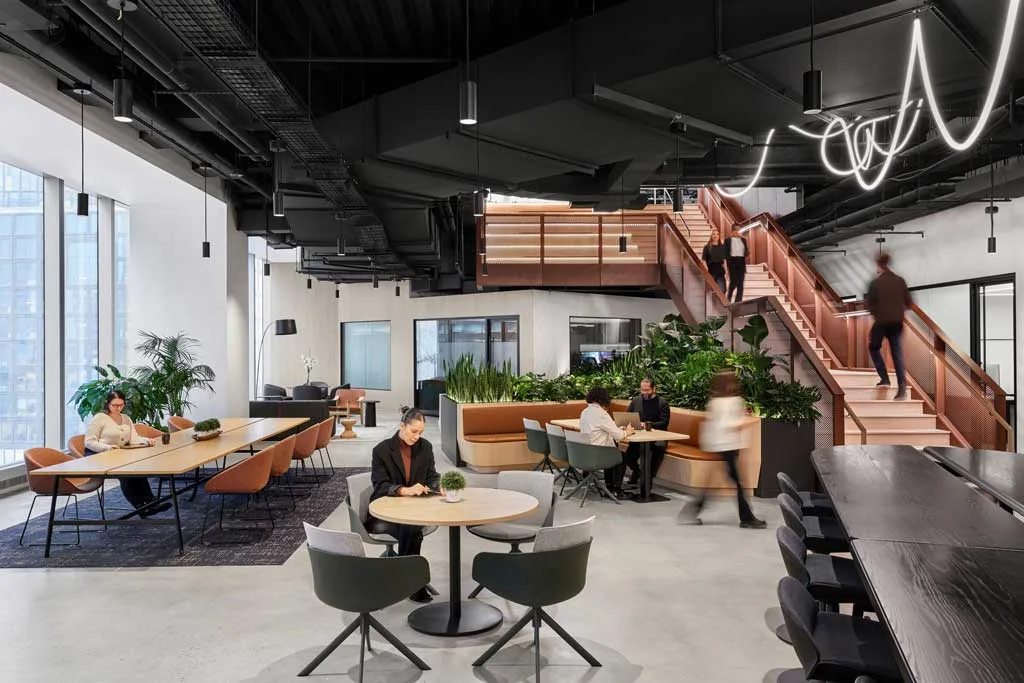 Area open space con tavoli condivisi, sedute moderne, scala scenografica in metallo color rame e illuminazione a sospensione presso l'HSBC Zero Carbon HQ sostenibile.