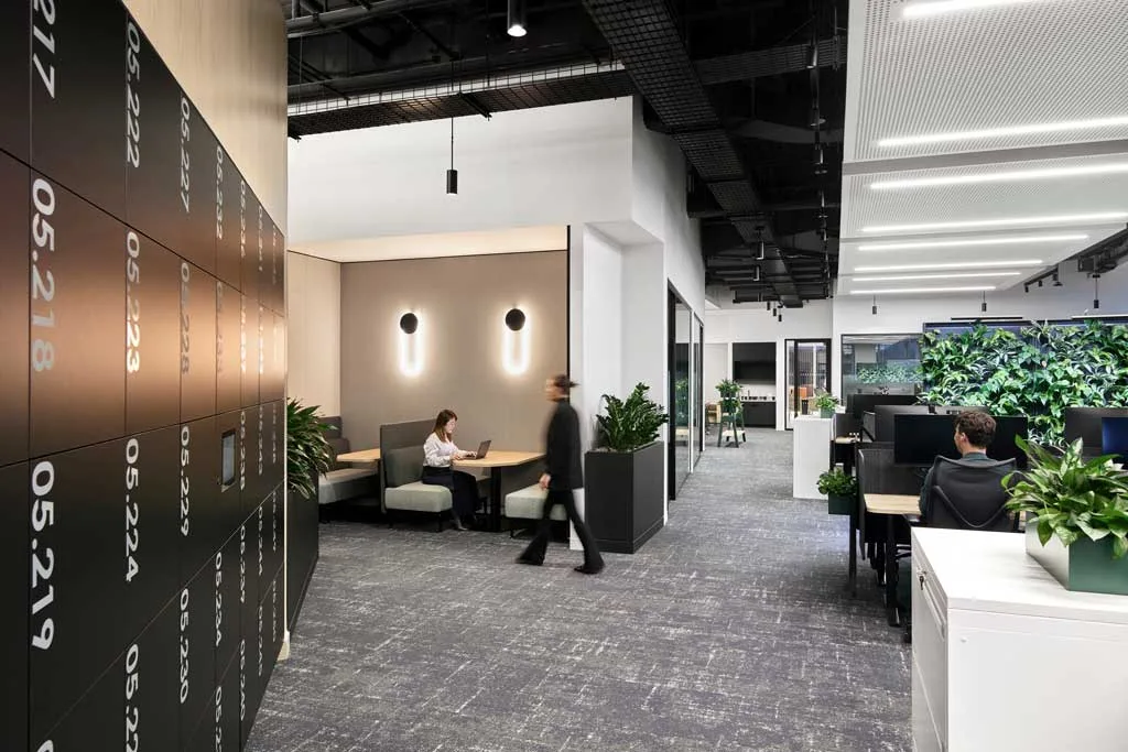 Area uffici open space con postazioni operative, parete verde, armadietti numerati e zona meeting informale con sedute imbottite presso l'HSBC Zero Carbon HQ sostenibile.