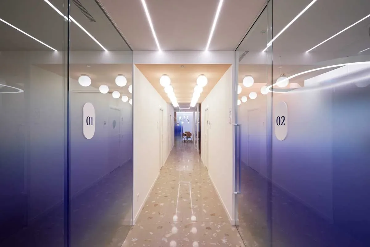 Corridoio centrale Visage Clinic con illuminazione a soffitto, pavimento effetto terrazzo e pareti in vetro blu sfumato.