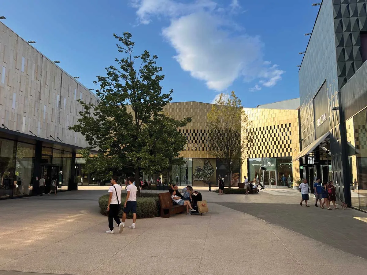 Piazza del San Marino Outlet Experience con facciate dorate, aree verdi e flusso di visitatori durante lo shopping.
