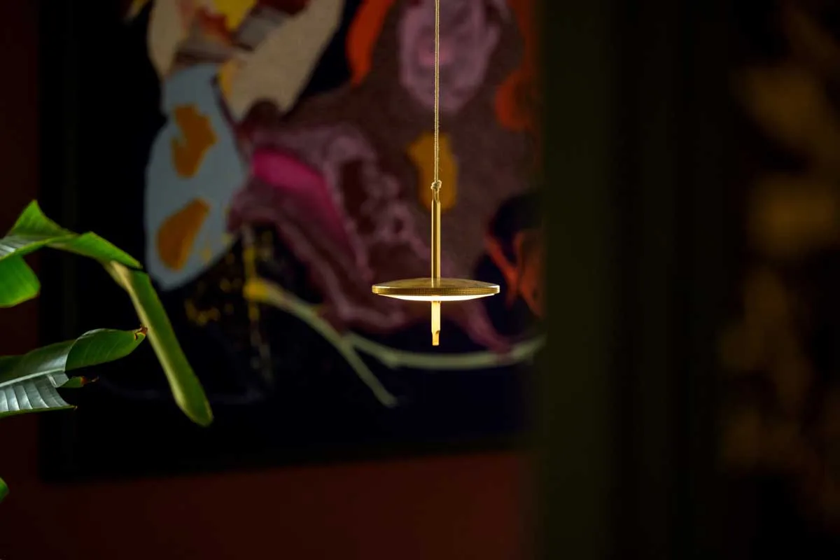 Lampada Thaïs di Platek sospesa con struttura in ottone davanti a parete decorata con opera astratta multicolore