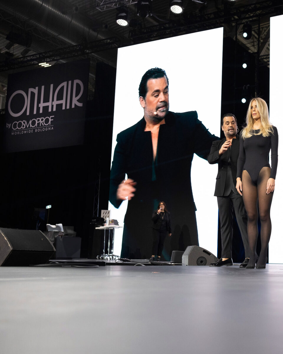 On Hair Show a Cosmoprof Bologna: hairstylist sul palco con modella e maxi schermo durante la presentazione