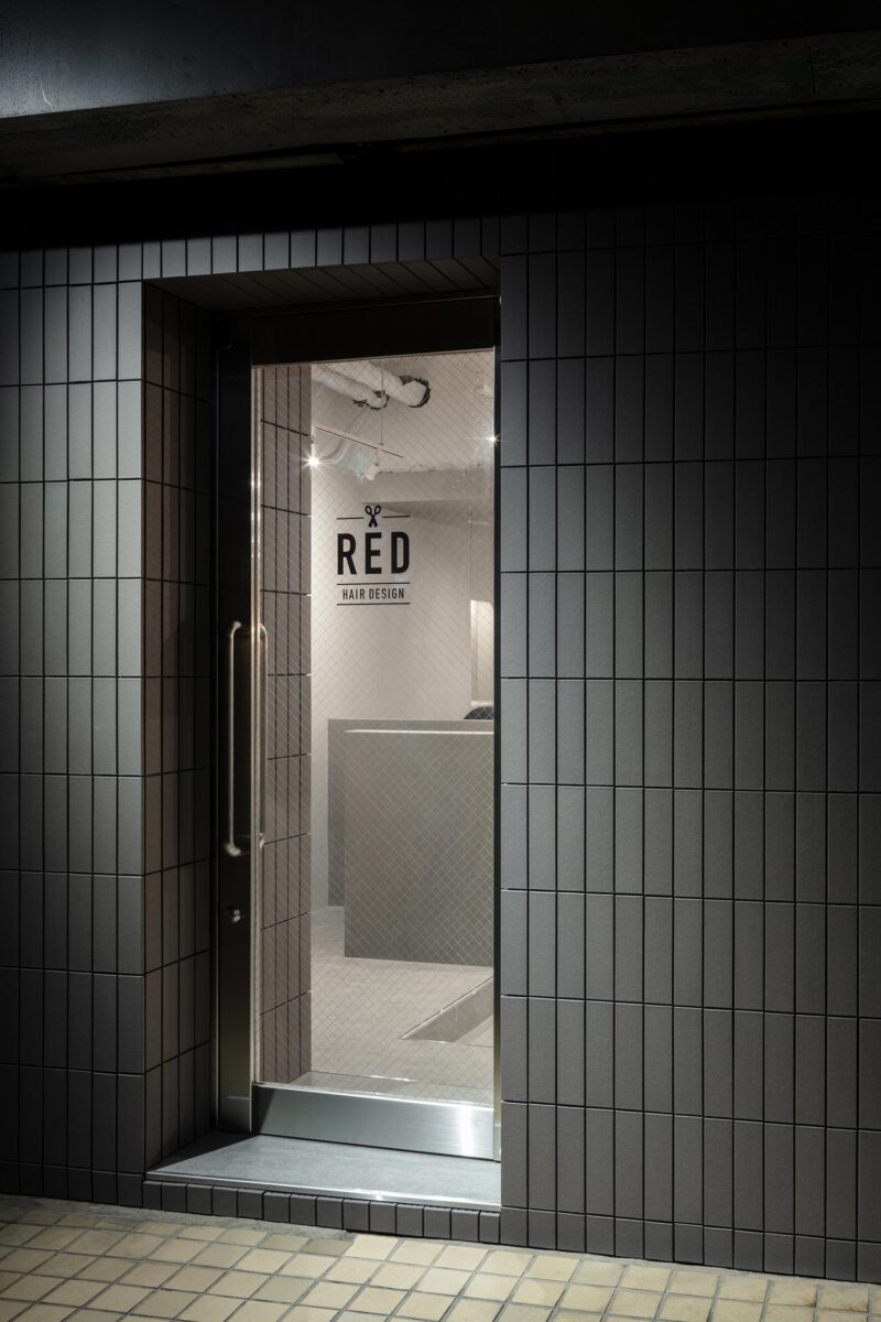 Entrance of RED Hair Design in Tokyo with dark tiled faรงade and glass door leading into a minimalist hair salon interior