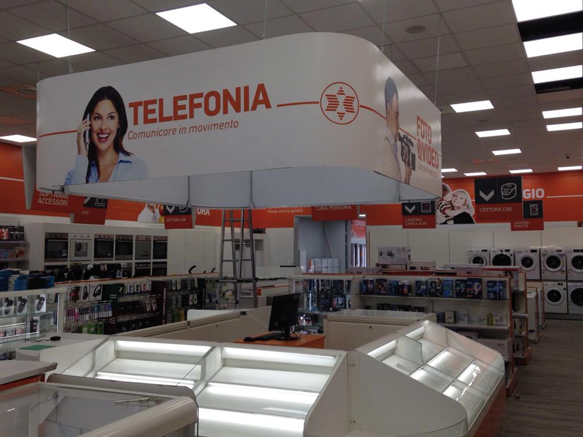 Electronics store interior with dedicated mobile phone area, illuminated display counter, suspended promotional signage, and household appliances department in the background.