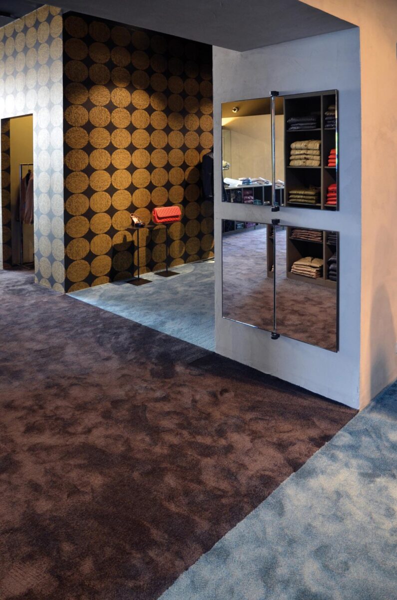 Boutique interior with two-tone textile flooring, decorative wall with gold circular pattern on dark background, integrated display niches, and open shelving for folded garments.