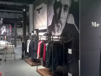 Interno di negozio di abbigliamento con arredi in metallo nero e legno, esposizione su rack lineari, grandi immagini fotografiche in bianco e nero alle pareti e illuminazione a binario.