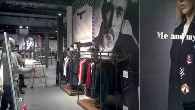 Interno di negozio di abbigliamento con arredi in metallo nero e legno, esposizione su rack lineari, grandi immagini fotografiche in bianco e nero alle pareti e illuminazione a binario.