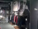 Interno di negozio di abbigliamento con arredi in metallo nero e legno, esposizione su rack lineari, grandi immagini fotografiche in bianco e nero alle pareti e illuminazione a binario.