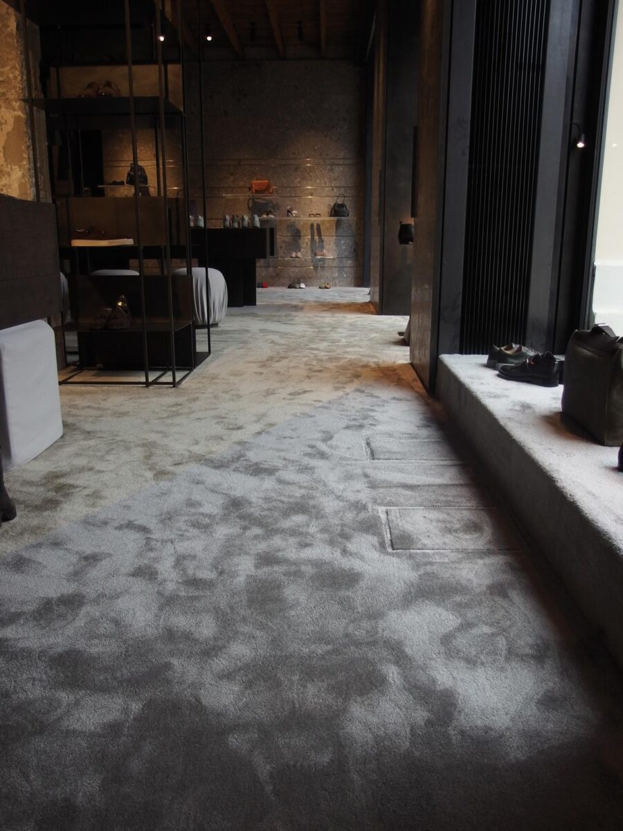 Boutique interior with light textile flooring, textured wall finishes, black metal and wood display fixtures, warm accent lighting, and accessories arranged on shelves and platforms.