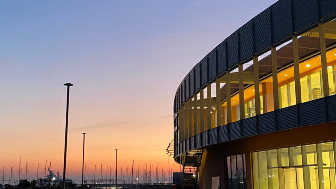 Vista esterna del Waterfront Mall di Genova al tramonto con facciata vetrata e affaccio sul porto