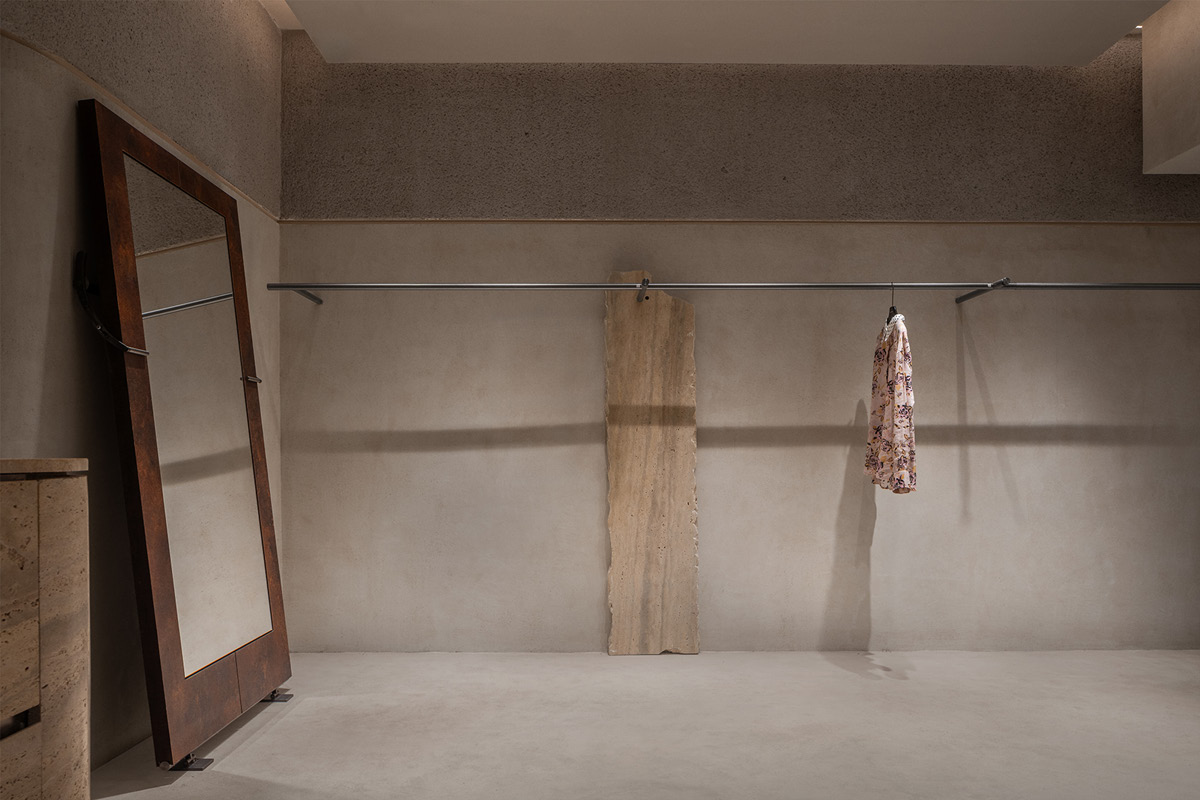 Minimal display system in the Hé showroom with a garment rail, full-height mirror and travertine slab
